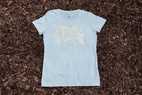 Light blue women's t-shirt with owl and oak design on top of mulch